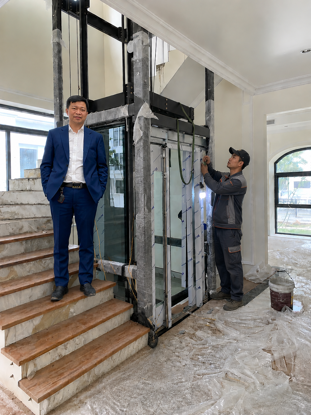Lắp đặt thang máy gia đình khung kính trong nhà, kỹ thuật viên thi công và khách hàng đứng cạnh thang máy